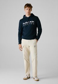 Marineblauwe hoodie met wit logo "Red Bull Racing", gecombineerd met crèmekleurige cargobroek met zijzakken en een gestructureerde afwerking. Sneakers met bruine accenten.