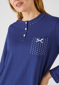 Haut à manches longues bleu marine avec un col rond, des boutons blancs décoratifs et une poche à pois avec un nœud en accent. Texture lisse.