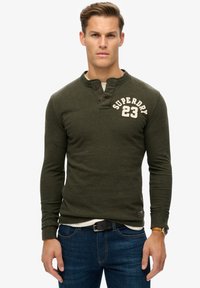Oliven grøn langærmet skjorte med en delt henley-krave, to knapper, hvid broderet "SUPERDRY 23" logo, båret over en hvid skjorte, parret med blå jeans.