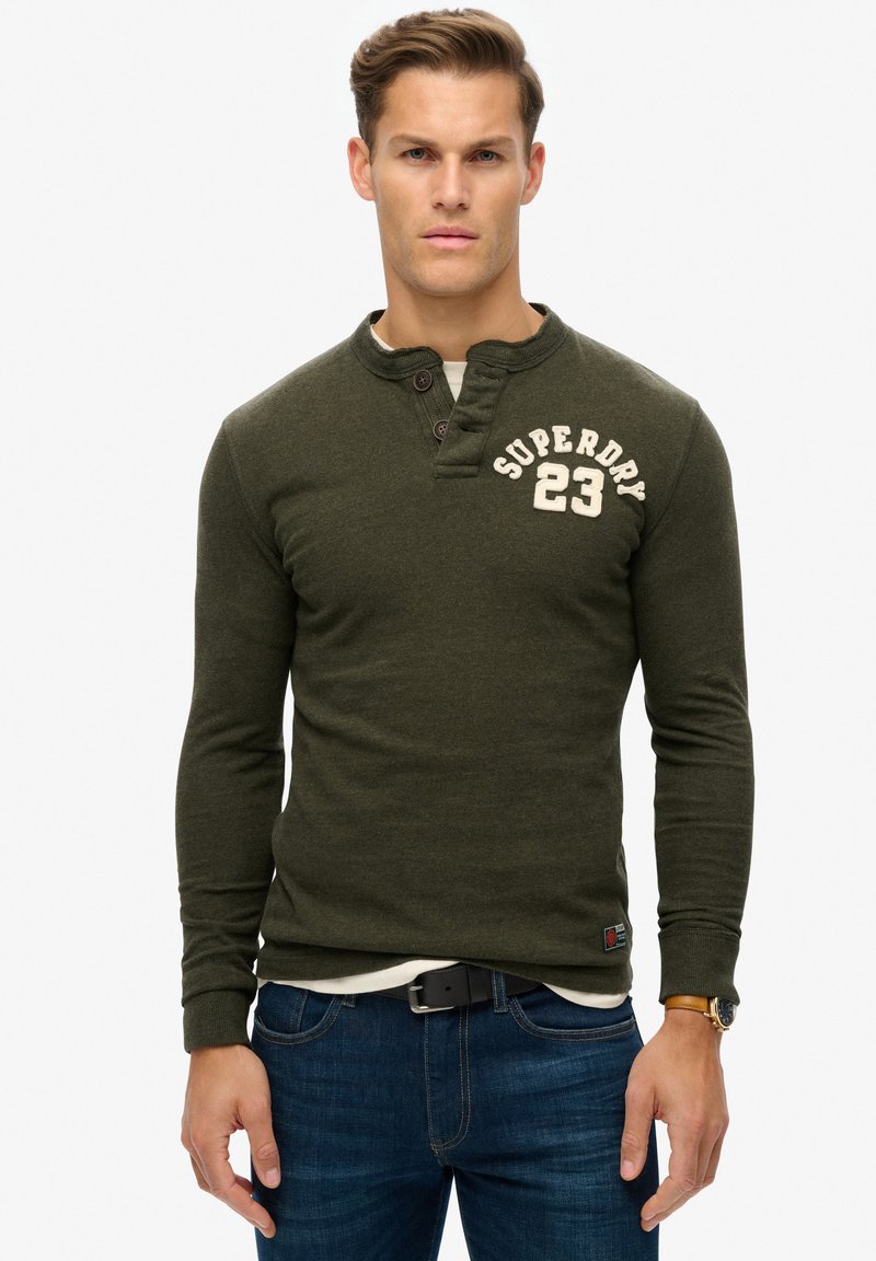 Oliven grøn langærmet skjorte med en delt henley-krave, to knapper, hvid broderet "SUPERDRY 23" logo, båret over en hvid skjorte, parret med blå jeans.