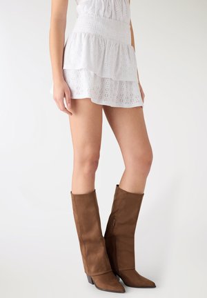 Donna che indossa una gonna corta bianca in pizzo a occhielli e stivali marroni di camoscio alti fino al ginocchio con tacchi a blocco, in piedi davanti a uno sfondo neutro.