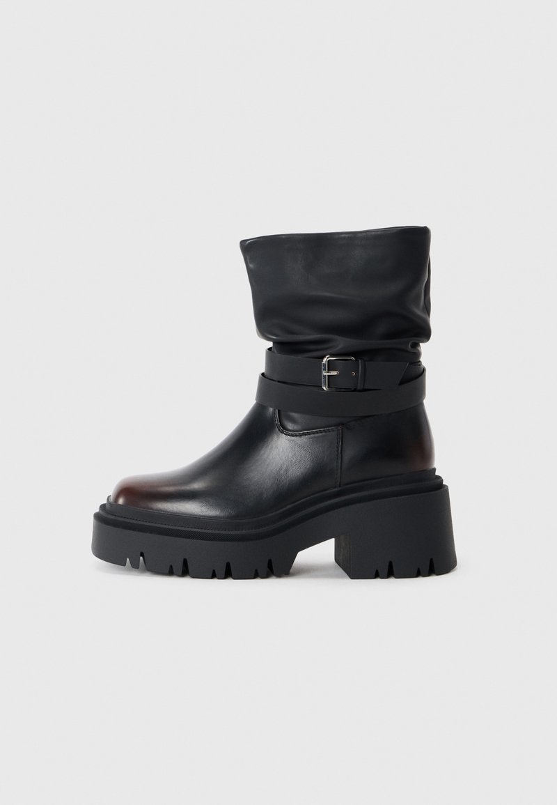 Bottine en cuir noir avec revers roulé, dotée d'une large sangle avec boucle, d'une semelle épaisse texturée à crampons profonds, et d'un talon épais.