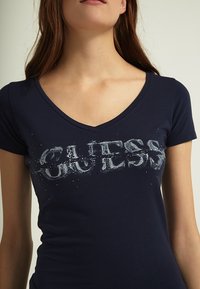 Marineblauwe V-hals t-shirt van katoen, uitgerust met een verweerd "GUESS" logo met zilveren accenten en kleine rhinestones aan de voorkant.