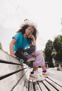 Zwarte rolschaatsen met kleurrijke wielen staan naast witte hoge sneakers met paarse en oranje accenten, op een houten bank.