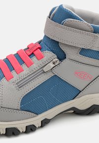 Pilki ir mėlyni žygių batai su rožiniais elastingais raišteliais, tekstūruota medžiaga, šoninė užtrauktukas ir tvirta juoda padas geresniam sukibimui.