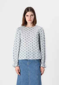 Blusa azul clara com padrão floral, mangas longas com punhos de babados e decote redondo. Usada com uma saia de ganga.