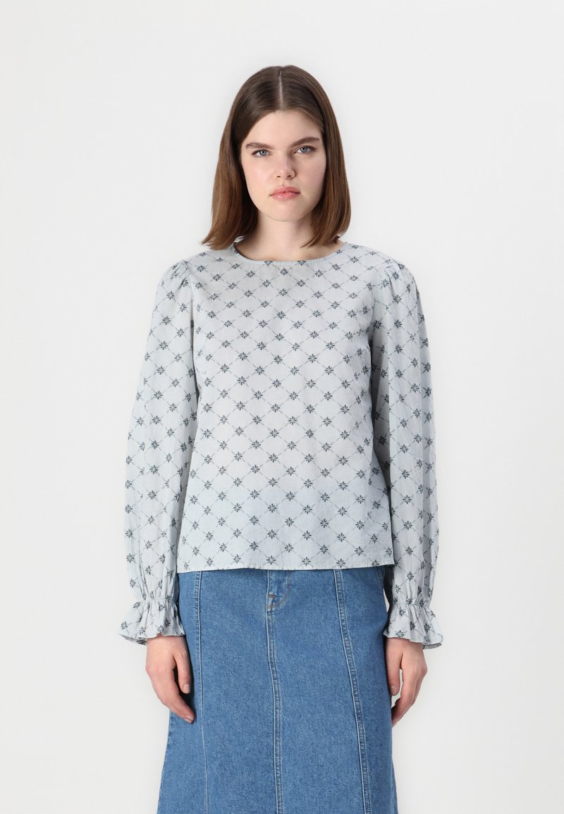 Blusa azul clara com padrão floral, mangas longas com punhos de babados e decote redondo. Usada com uma saia de ganga.