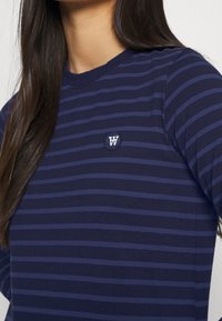 Granatowa koszula z długim rękawem z ciemniejszymi niebieskimi poziomymi paskami oraz małym okrągłym emblematem z literą "W" na piersi. Gładka tekstura materiału.