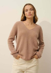 Jasnobrązowy sweter w serek z żebrowanym wzorem i długimi rękawami; zestawiony z kremowymi spodniami, tworząc miękki kontrast kolorystyczny.