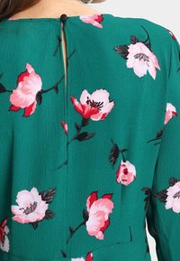 Zelená texturovaná látka s růžovým a červeným květinovým vzorem, s výstřihem ve tvaru klíčové dírky, zapnutým černým knoflíkem na horní části zad.