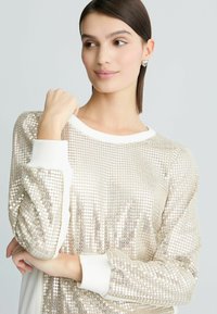 Top lungo a maniche con paillettes dorate lucide, con polsini e scollatura bianchi; presenta una vestibilità morbida e una superficie texture.