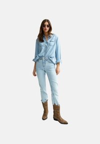 Camicia di jeans blu chiaro, jeans blu chiaro e stivali da cowboy marroni con cuciture decorative. Modello che indossa occhiali da sole tondi. Sfondo neutro.