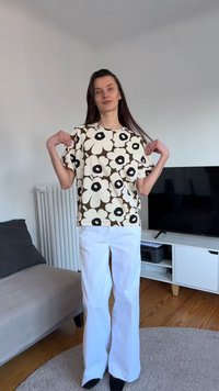 Γυναίκα μέσα στο σπίτι που φοράει άσπρο πουκάμισο με λουλουδάτο μοτίβο και άσπρο παντελόνι, κρατώντας τον γιακά του πουκαμίσου με τα δύο χέρια, στέκεται κοντά σε έναν καναπέ και τηλεόραση.