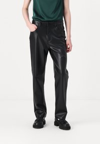 Zwarte faux leren broek met een rechte pasvorm, voorzakken en zichtbare stikdetails. Outfit gestyled met een donkergroen topje en zwarte schoenen.