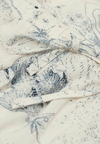 Baumwollstoff mit einer hellcremefarbenen Basis, der mit einem blauen Druck verschnörkelter Designs und einer gebundenen Detail in den Stofffalten versehen ist.