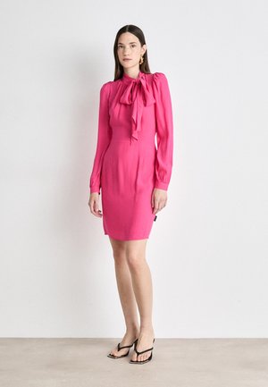 Fuchsia dress with long sleeves, high neckline, and bow detail. Smooth fabric, fitted silhouette, reaching mid-thigh. Black strappy sandals.