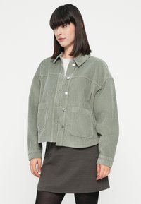 Donna con capelli neri e lisci che indossa una giacca di velluto a coste verde chiaro, una gonna a quadretti e collant neri su uno sfondo bianco.
