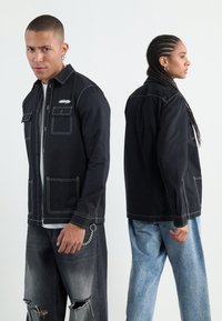 Schwarze Button-Up-Jacke mit kontrastierenden weißen Nähten, zwei Brusttaschen und einem lässigen Schnitt, kombiniert mit verwaschenen grauen Jeans.