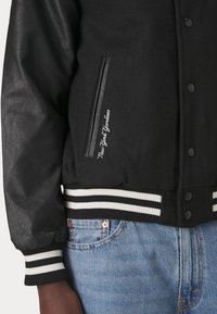 Svart collegejacka med läderärmar, randiga ribbade manschetter och nederkant, broderi av "New York Yankees", bärs med blåjeans.