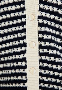 Navy and ivory striped cardigan with textured horizontal lines, featuring a row of six button closures made of a light-colored material.