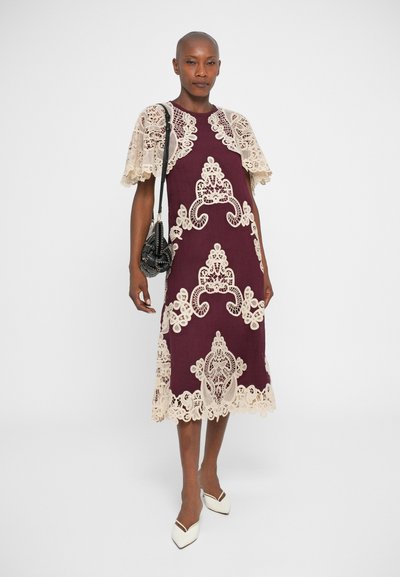 Vestido bordô com detalhes em renda, com mangas curtas e padrões; acabamentos e recortes em renda creme detalhados; combinado com uma pequena bolsa preta.