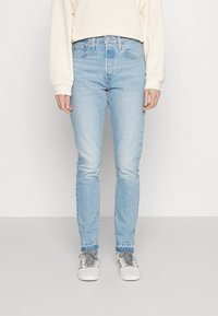 Osoba v lehkých modrých slim-fit džínách, béžové mikině a bílých teniskách se šedými tkaničkami, stojí na jednobarevném pozadí.