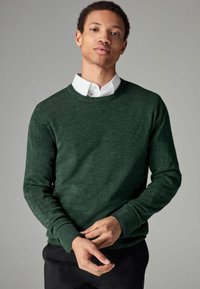Suéter de punto verde oscuro con cuello redondo, mangas largas y puños de canalé, llevado sobre una camisa blanca con cuello, de textura de tela ligera.