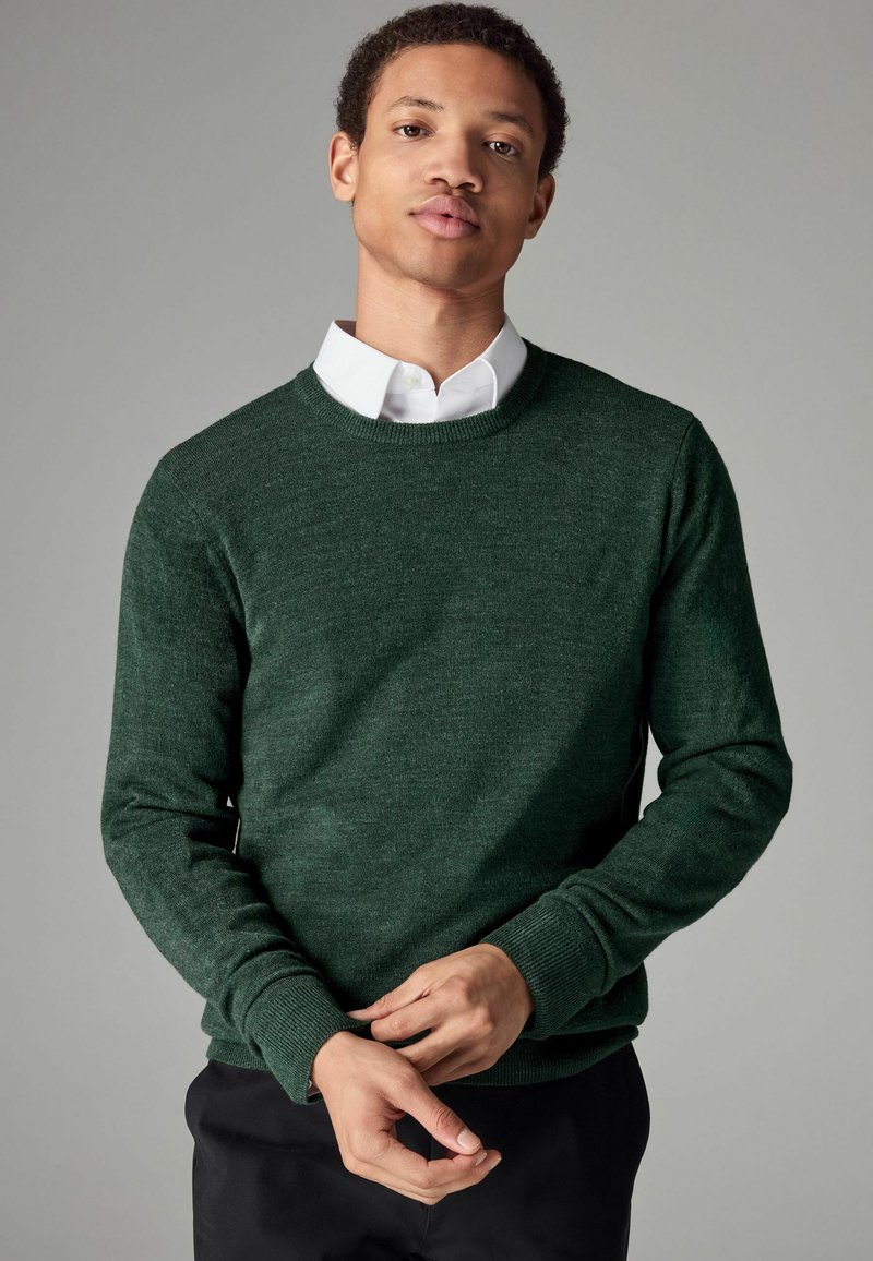 Suéter de punto verde oscuro con cuello redondo, mangas largas y puños de canalé, llevado sobre una camisa blanca con cuello, de textura de tela ligera.