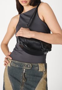 Svart lærveske med buet form og nagler, båret over kroppen. Modellen har på seg en grå ermeløs topp og et denimskjørt med brune detaljer.