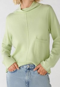 Donna che indossa un maglione a maglia verde chiaro con tasca frontale e jeans di denim blu chiaro, mani in tasca su uno sfondo semplice.