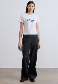 Λευκό βαμβακερό t-shirt με μπλε λογότυπο "CALVIN", συνδυασμένο με μαύρο τζιν παντελόνι φαρδιάς γραμμής. Το μοντέλο κρατάει μια ασημί μεταλλική τσάντα χειρός.