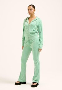 Tuta in velluto verde menta con giacca a zip e hardware dorato, tasche frontali e pantaloni svasati. Finitura in tessuto morbido e dal texture piacevole.