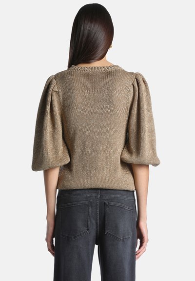 Femme aux longs cheveux foncés portant un haut en maille métallique dorée avec des manches bouffantes jusqu'aux coudes et un jean noir en denim, vue de dos.