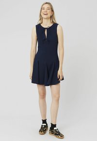 Robe sans manches bleu marine avec un décolleté en trou de serrure et un détail de nouage, jupe plissée évasée, associée à des baskets à imprimé léopard et des chaussettes noires.