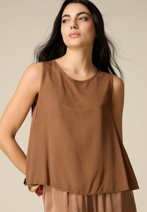 Donna con lunghi capelli scuri che indossa una canotta marrone, guarda leggermente di lato su uno sfondo beige uniforme.