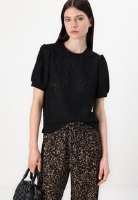 Zwarte gestructureerde gebreide top met korte pofmouwen, gecombineerd met goud- en bruingekleurde luipaardprint broek. Model houdt een kleine zwarte handtas vast.
