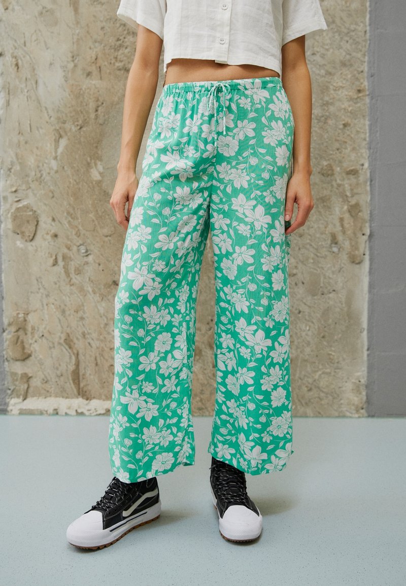 Pantalons larges floraux en tissu teal, avec un motif floral blanc, taille élastique et poches latérales, associés à des baskets noires.