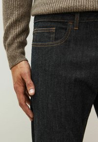Prim-plan al unei persoane îmbrăcate cu blugi închiși la culoare și un pulover tricotat maro, cu mâna stângă odihnindu-se lângă coapsă.