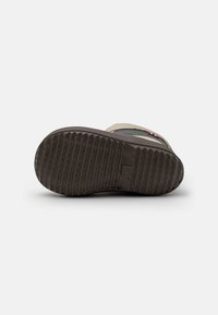 Semelle de botte en caoutchouc, texturée avec des rainures profondes. Couleur : marron foncé. Caractéristiques d'un bord surélevé et de détails de logo. Bord supérieur lisse avec des accents beige clair.