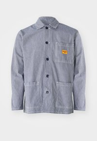 Striped work jacket in navy and white, featuring a button-up front, collar, chest pocket with yellow label, and two front pockets.