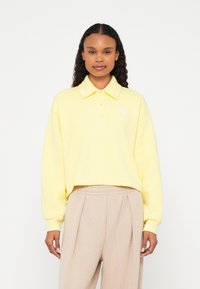 Polo cropped amarelo com textura canelada, pala com dois botões e um pequeno detalhe em forma de diamante. Combinado com calças plissadas bege.