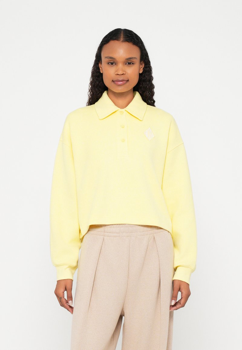 Polo cropped amarelo com textura canelada, pala com dois botões e um pequeno detalhe em forma de diamante. Combinado com calças plissadas bege.