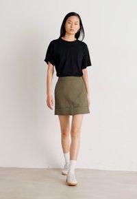 Svart t-skjorte med korte ermer, olivengrønn tettsittende skjørt, hvite ankelsokker og beige snøresko på en modell foran en ensfarget bakgrunn.