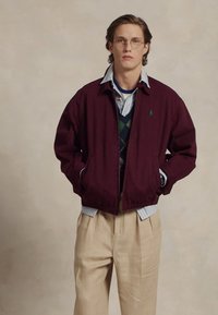 Bordeaux bomberjack met een gestructureerde vorm, gedragen over een groen-blauw geruite trui en een lichtblauwe gestreepte shirt, gecombineerd met beige broeken.