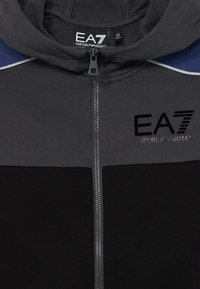Čierna a tmavo šedá bunda s kapucňou na zips s modrými akcentmi, logo EA7 Emporio Armani na hrudi a viditeľná veľkostná etiketa vo vnútri goliera.