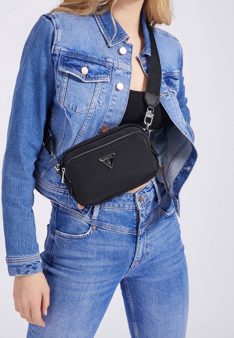 Schwarze Umhängetasche mit rechteckiger Form, ausgestattet mit einem Reißverschluss und einer Metall-Logoplatte. Getragen über einer Jeansjacke und Jeans.