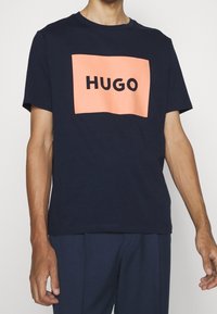 Námořnické bavlněné tričko s obdélníkovou broskvovou grafikou, na které je v tučných černých písmenech napsáno "HUGO". Krátké rukávy, design s kulatým výstřihem.