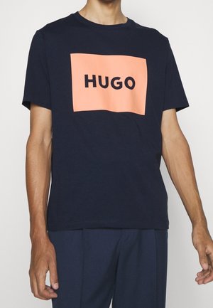 Marineblaue Baumwoll-T-Shirt mit einer rechteckigen, pfirsichfarbenen Grafik, auf der "HUGO" in fetten schwarzen Buchstaben steht. Kurzärmlig, Rundhalsausschnitt.