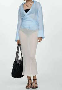 Camicia avvolgente blu chiaro con maniche lunghe, abbinata a una gonna bianca trasparente. Il modello tiene una borsa nera intrecciata e indossa sandali neri con lacci.