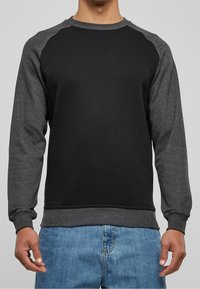 Homme portant un sweat-shirt à manches longues noir et gris foncé associé à un jean en denim bleu, debout devant un fond gris uni.